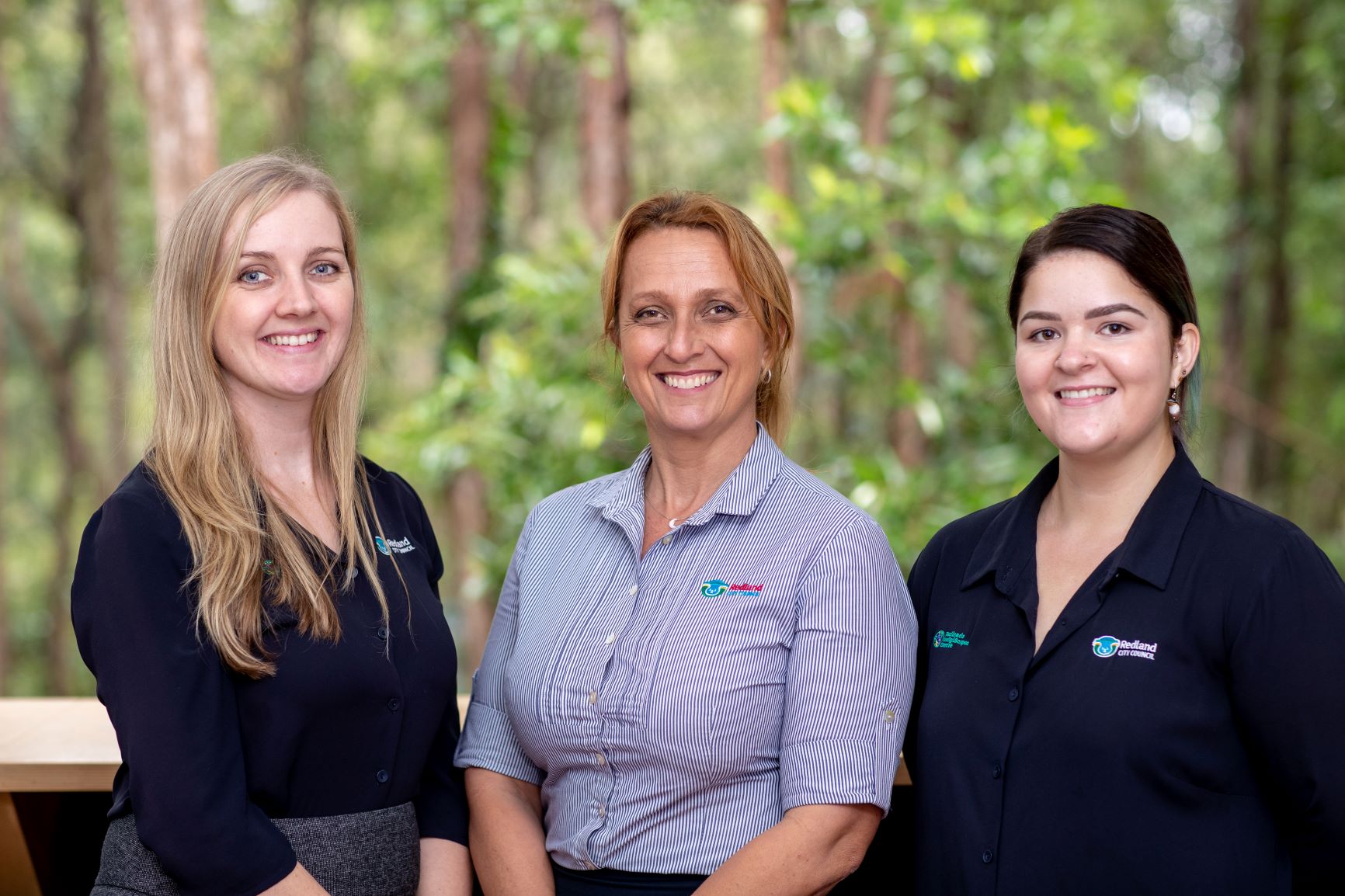 Three women standing together ready to serve you with bushland in the background.