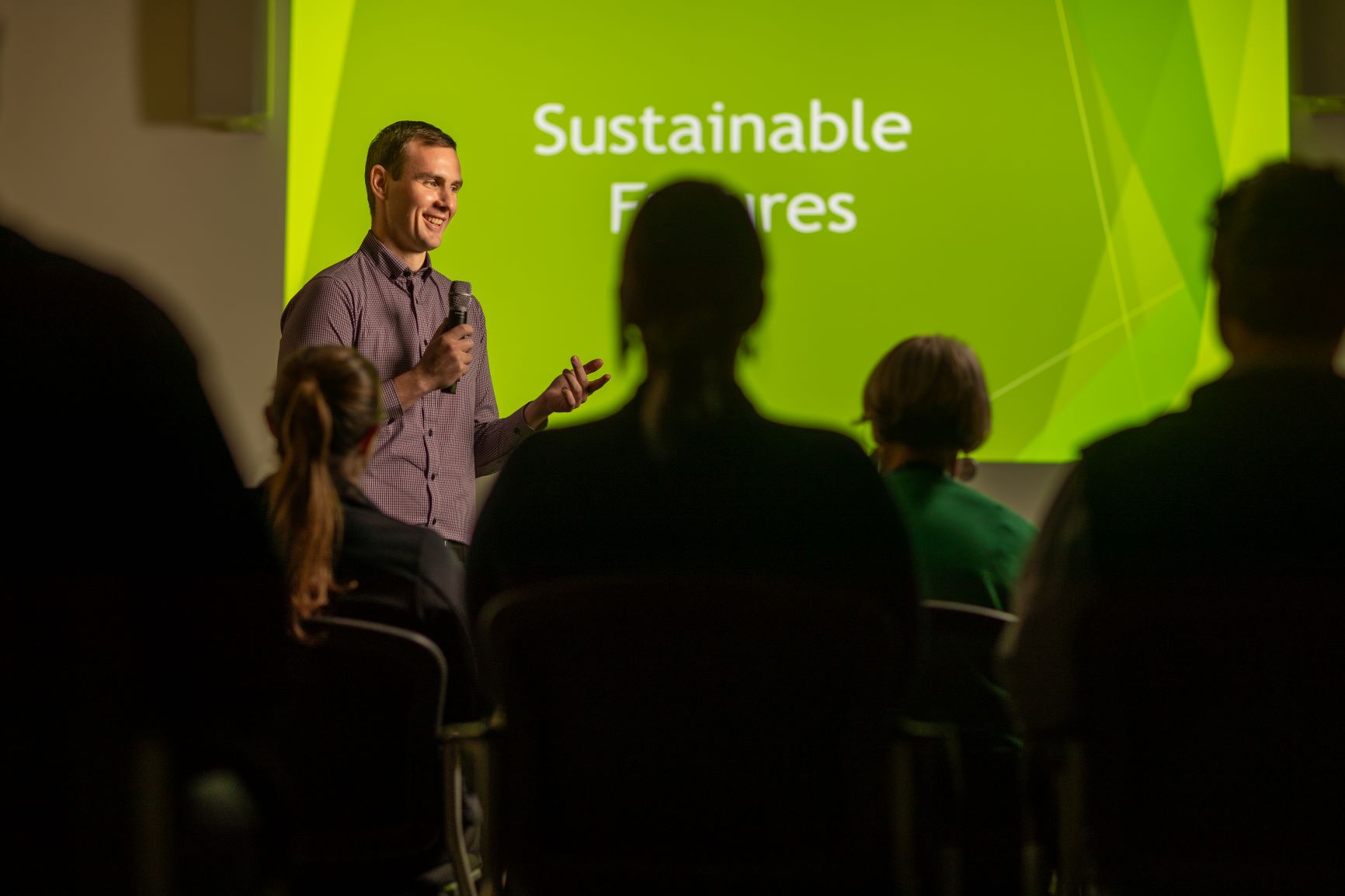 Man with a microphone standing in front of a large screen with the words Sustainable Features and people in the audience listening.